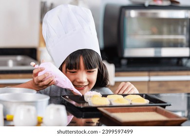 Closeup shot of Asian female little girl baker pastry bakery chef daughter wears white tall hat standing smiling using colorful whipped cream decorating preparing homemade cupcakes in home kitchen. - Powered by Shutterstock