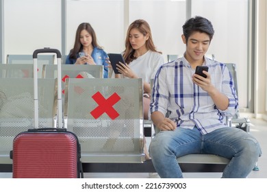 Closeup Shot Of Asian Male Passenger Sitting Browsing Surfing Internet Via Smartphone On New Normal Social Distancing Chairs With Red Cross Sticker In Airport During Covid19 Pandemic Quarantine.