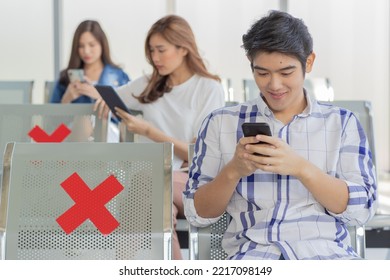 Closeup Shot Of Asian Male Passenger Sitting Browsing Surfing Internet Via Smartphone On New Normal Social Distancing Chairs With Red Cross Sticker In Airport During Covid19 Pandemic Quarantine.