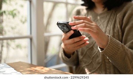 Close-up shot of an Asian woman in cozy sweater using her smartphone, chatting with someone or scrolling on her phone while relaxing at a cafe. - Shutterstock ID 2299629245