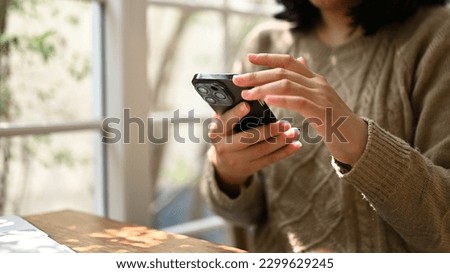 Close-up shot of an Asian woman in cozy sweater using her smartphone, chatting with someone or scrolling on her phone while relaxing at a cafe.
