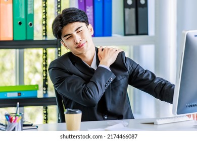 Closeup shot of Asian young tired exhausted frowning face male businessman employee staff in formal suit sitting at working desk take break holding hands on back has painful backache office syndrome.
