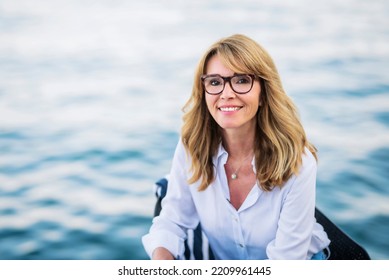 Close-up Shot Of Attractive Mature Woman Wearing White Shirt And Blue Jeans While Sitting On The Chair And Relaxing By The Sea.