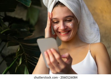 Close-up Shot Of A Beautiful Young Woman Doing Her Morning Routine, Applying Face Cream While Covering Her Hair With A Towel After Washing And Texting On The Phone At The Same Time.