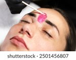 A close-up shot of a beauty professional applying pink wax between a woman’s eyebrows. The woman’s eyes are closed, focusing on the precise application of the wax for facial grooming.