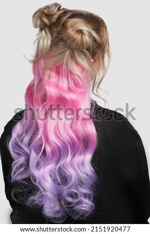 Close-up shot of a blonde girl with curly pink and purple strands of hair. Ombre clipin hair extensions. The woman in the black sweater with tresses is on a gray background. Back view.