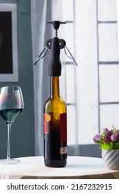 Close-up Shot Of A Bottle Of Red Wine With A Black Winged Corkscrew. The Bottle Of Red Wine With A Corkscrew, An Empty Glass And A Vase Of Flowers Are On The Table In The Gray Room. Front View.