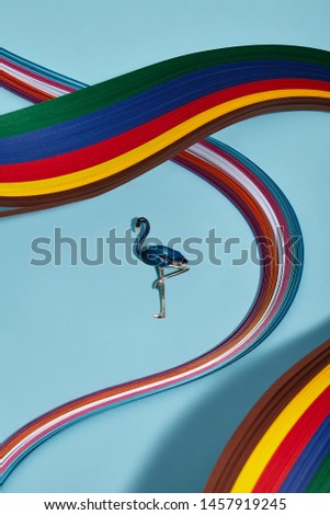 Closeup shot of breastpin in view of flamingo with navy blue feathers, isolated asymmetrically on light blue background in midst of multicolored wavy placed ribbons. Fashionable women's fashion item.