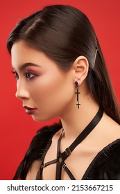 Close-up shot of a brown haired girl with black earrings with two cross pendants. Stud earrings are in the gothic style. The woman with earrings is posing on a red background. Side view. - Shutterstock ID 2153667215