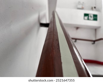 A close-up shot of a brown handrail with a white stripe running down the center. The handrail is mounted to a white wall. The background is blurred and shows a red emergency exit sign - Powered by Shutterstock