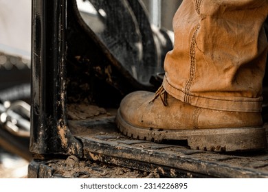A close-up shot of brown leather boot of a construction worker sitting on an aged dirty tractor in a rural setting
