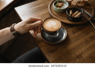 A close-up shot of a cappuccino cup with a heart-shaped foam in the hands of a man, enjoying his breakfast in a cozy cafe. - Powered by Shutterstock