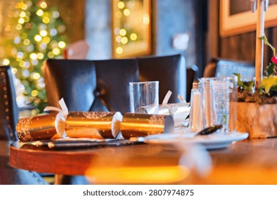 A close-up shot captures an elegant table setting during a Christmas event, with crisp focus on the glassware and folded napkins. The warm, low-key lighting creates a festive and intimate mood, dramatically enhanced by the blurred bokeh of a decorated Christmas tree in the background.