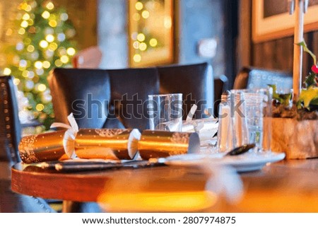 A close-up shot captures an elegant table setting during a Christmas event, with crisp focus on the glassware and folded napkins. The warm, low-key lighting creates a festive and intimate mood, dramatically enhanced by the blurred bokeh of a decorated Christmas tree in the background.