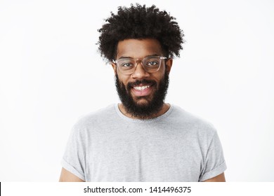 Close-up shot of charming pleasant and delighted dark-skinned bearded male customer in glasses with afro hairstyle in grey casual t-shirt smiling joyfully as having great mood over gray background