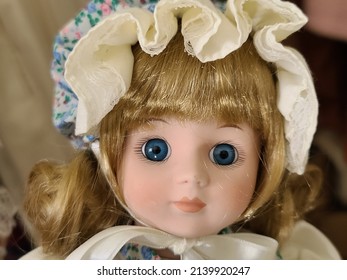 A closeup shot of a china doll's face. The doll is wearing a frilly bonnet with a floral design, has blonde hair, and bright blue eyes.