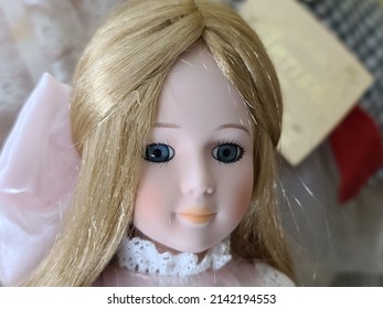 A Closeup Shot Of A China Doll's Face. This Porcelain Doll Has Long Blonde Hair, Blue Eyes, And A Giant, Pink Bow Holding Her Hair Back.