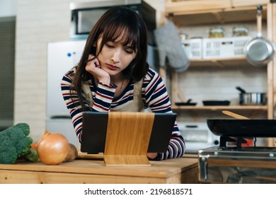 Closeup Shot Of A Confused Asian Female Kitchen Novice Propping Her Face And Leaning Near The Electric Pad While Reading Online Recipes To Learn Cook At Home.