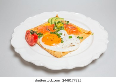 Close-up shot of a crepe with smoked salmon, avocado, egg, and tomatoes on a white plate. Square shape, elegant presentation.