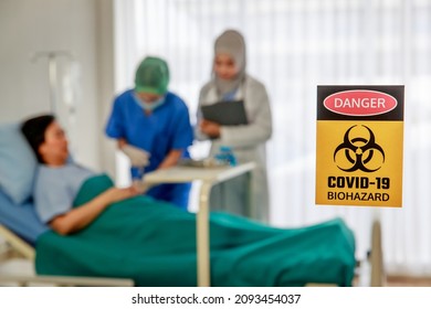 Closeup Shot Danger Biohazard Covid-19 Yellow Hazard Sign Symbol Banner In Front Blurred Background Of Muslim Islam Arab Doctor Visit Patient While Nurse Inject Saline Solution In Hospital Ward Room.