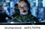 Close-up shot of Female Army Officer Using Computer in Headphones. In the Background Busy System Control Center with People Working, Displays with Information.