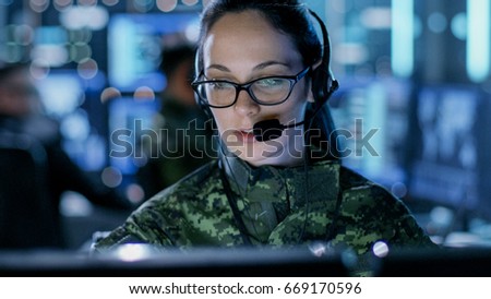 Close-up shot of Female Army Officer Using Computer in Headphones. In the Background Busy System Control Center with People Working, Displays with Information.