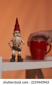 Close-up Shot Of A Female Hand Holding A Red Cup. The Figurine Of A Skeleton Of A Dwarf Man In A Red Hat And A Brown Loincloth Is Located On A Bookshelf On The Orange Background.                      