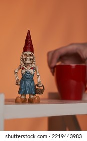 Close-up Shot Of A Female Hand Holding A Red Cup. The Figurine Of A Skeleton Of A Dwarf Woman In A Red Hat And A Blue Dress With A Basket In Her Hand Is Located On A Bookshelf On An Orange Background