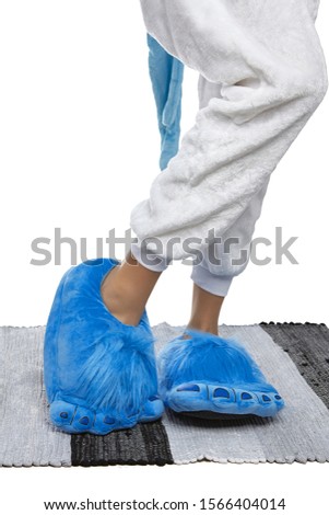 Close-up shot of female legs in white velour trousers and blue plush house slippers made as shaggy hobbit feet. The girl is standing on the striped gray and white carpet.