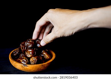 A close-up shot of a hand selecting a date from a small wooden bowl filled with sweet, brown dates, set against a stark black background, highlighting the rich texture and color of the fruit.