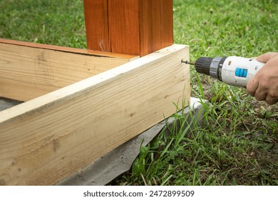 A close-up shot of a hand using a cordless drill to attach a wooden beam to a concrete foundation. The beam is likely part of a deck or pergola. - Powered by Shutterstock