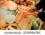 A close-up shot of hands shredding fresh carrots and cabbage at a market stall. Piles of shredded vegetables and whole zucchinis display the fresh, organic produce ready for cooking