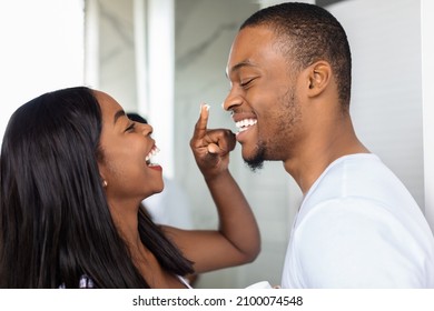 Closeup Shot Of Happy African American Couple Having Fun In Bathroom, Cheerful Black Woman Applying Moisturising Cream On Husband's Face, Playfully Touching His Nose And Laughing Together
