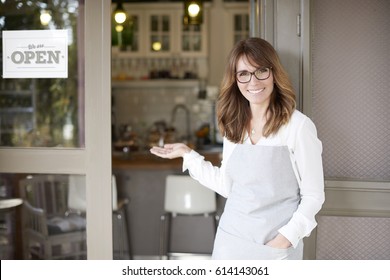 Close-up shot of a happy small business owner standing at doorway and inviting customers while looking at camera and smiling. 