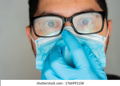 A Closeup Shot Of A Hispanic Man Wearing A Blue Disposable Face Mask, Foggy Glasses, And Plastic Gloves, Looking At The Camera