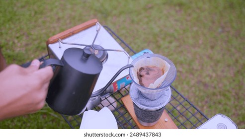 A close-up shot of hot water being poured into a dripper to brew fresh coffee outdoors on a foggy morning with a scenic green background at camping campsite - Powered by Shutterstock