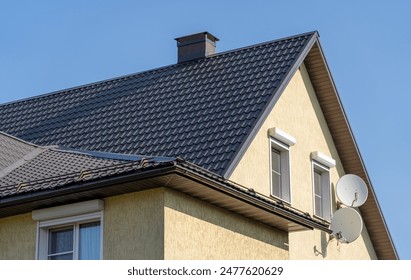 Close-up shot of a house with a black metal roof, a chimney, and two windows on the side. The house is painted a pale yellow