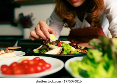 Closeup Shot Of Houswife Hand Decorating Chicken Breast Green Mixed Vegetable Salad With Boiled Egg Yolk On Countertop Desk In Kitchen With Raw Fresh Organic Material In Blurred Foreground.