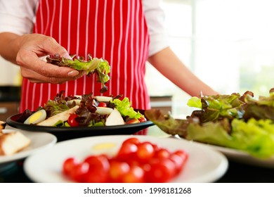 Closeup Shot Of Houswife Hand Decorating Chicken Breast Green Mixed Vegetable Salad With Boiled Egg Yolk On Countertop Desk In Kitchen With Raw Fresh Organic Material In Blurred Foreground.