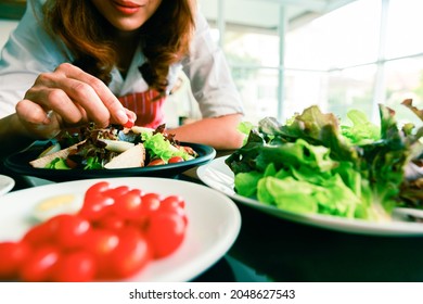 Closeup Shot Of Houswife Hand Decorating Chicken Breast Green Mixed Vegetable Salad With Boiled Egg Yolk On Countertop Desk In Kitchen With Raw Fresh Organic Material In Blurred Foreground.