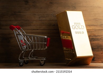 Closeup Shot Of A Large Shiny Gold Bar Near The Tiny Shopping Cart With Wooden Wall Background.