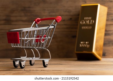 Closeup Shot Of A Large Shiny Gold Bar Near The Tiny Shopping Cart With Wooden Wall Background.