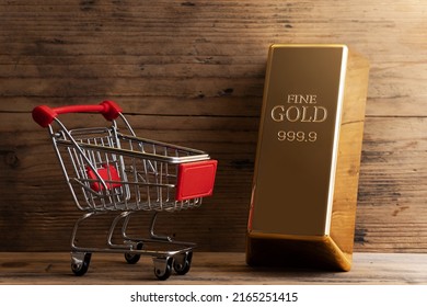 Closeup Shot Of A Large Shiny Gold Bar Near The Tiny Shopping Cart With Wooden Wall Background.