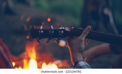 Close-up shot of male tourist's hand playing the guitar during romantic evening at campsite with fire burning in background. Musical instruments, nature and people concept.