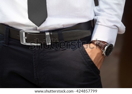 Closeup shot of male waist with hand in pocket dressed in black pants, belt, grey shirt, black tie and watch with brown watch strap. Formal wear.
