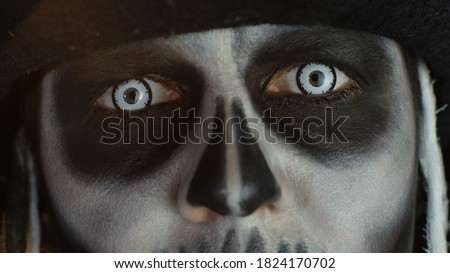 Close-up shot of man face in skeleton Halloween cosplay costume. Guy in creepy skull makeup opening eyes with white pupil, trying to scare. Day of The Dead. Isolated on black background