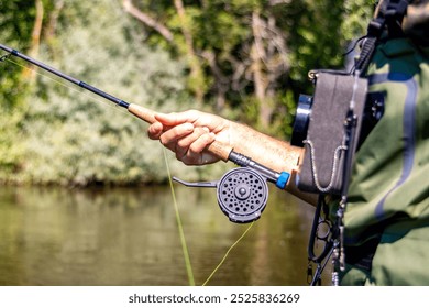 A close-up shot of a man holding a fly fishing rod near a river. The image captures the details of the fishing gear and the man's hand, emphasizing focus and outdoor activity. - Powered by Shutterstock