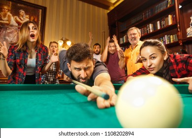 Close-up shot of a man playing billiard. The caucasian model carefully and strenuously aiming by cue in the ball. Game concept. Human emotions and facial expression concepts. Friends as background