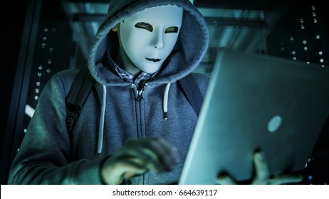 Close-up Shot of a Masked Hacker in a Hoodie Standing in the Middle of Data Center full of Rack Servers and Hacking it with His Laptop.