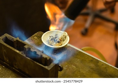 Closeup shot of a melting dish with metal rings and other silver pieces being heated with a torch flame. The intense heat causes the metal to begin melting, creating a molten base for jewelry casting. - Powered by Shutterstock